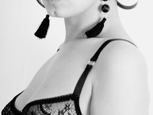 Le buste de Zazazelle, soutien-gorge en dentelle noire, glands dans les oreilles, bretelles sombres, vue de profil partielle, fond blanc, noir et blanc, lumière douce, clavicule visible, seins légèrement apparents..