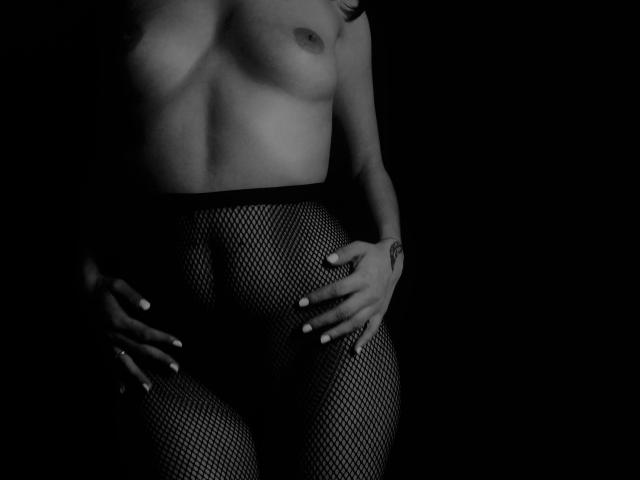Zazazelle est nue, avec de petites seins, elle porte un pantalon en résille noir, ses mains sont sur son entrejambe, l'arrière-plan est sombre, en noir et blanc, et on peut voir un tatouage à son poignet droit..