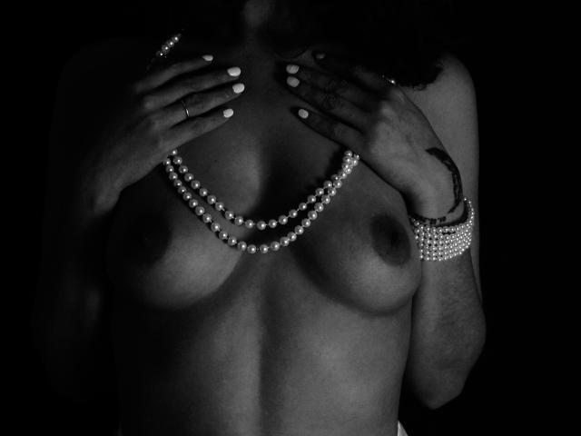 Zazazelle tiene la piel oscura, pechos pequeños y está sin sujetador, con las manos cubriéndolos. Lleva un collar de perlas y una pulsera, con uñas blancas y un tatuaje..