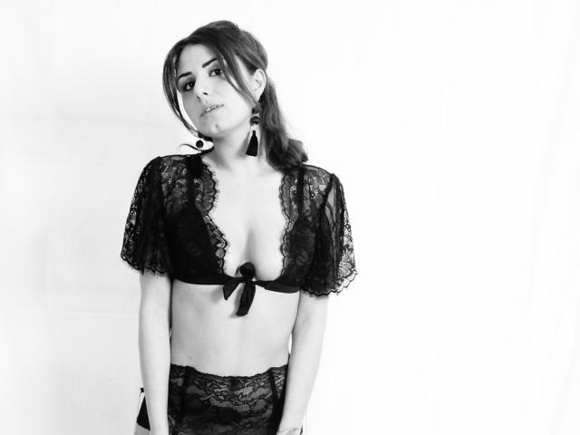 Zazazelle in biancheria intima di pizzo nero, con i seni appena coperti da un laccio anteriore, che guarda di lato con un leggero sbuffo, indossando orecchini lunghi, in una foto in bianco e nero..