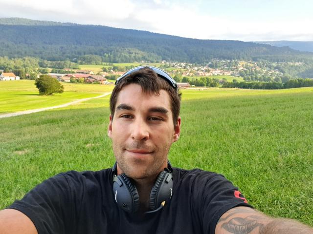 DarkPunkX tiene el pelo corto y castaño, una ligera sonrisa y auriculares negros alrededor del cuello, vistiendo una camiseta negra con un tatuaje en el brazo derecho, en un campo cubierto de hierba..