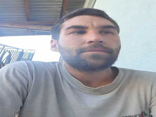 DarkPunkX, con una expresión ligeramente molesta, cabello castaño corto y barba, vistiendo una camisa gris, de pie debajo de un techo de metal, con una pared blanca en el fondo..
