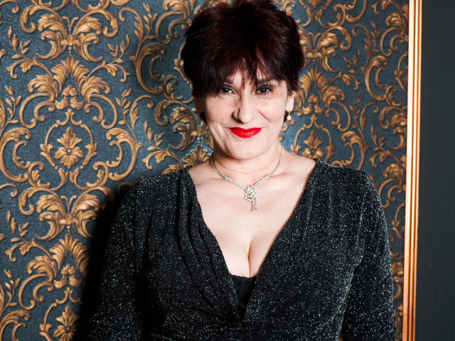 AlbertaAllen, short dark red hair, red lipstick, sparkling black dress, deep cleavage, necklace, smiling, dark blue and gold patterned wallpaper, gold framed mirror on the right.