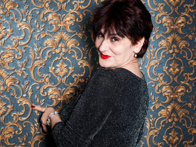 AlbertaAllen with short dark brown hair, red lipstick, black sparkly top, pointing with red-manicured fingers, smiling over shoulder, gold earrings, wristwatch, and confident look.