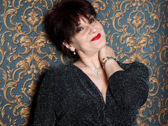 AlbertaAllen with short brown hair, red lipstick, sparkling black top, hand on neck, elegant bracelet, and necklace, smiling at camera, showing off her tits slightly.