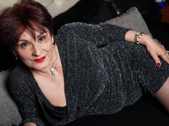 AlbertaAllen has short red hair, red lipstick, and sparkly black dress, lying on a black couch, hand on thigh, wearing a necklace and bracelet, looking at the camera.