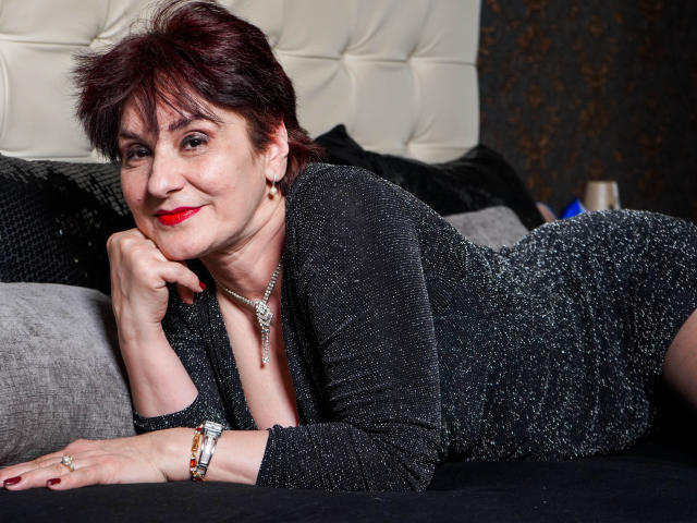 AlbertaAllen, middle-aged woman with short red hair, lying on bed, black sparkly dress, red lipstick, smiling, hand under chin, wearing bracelet and necklace, cute, attractive, ass visible.