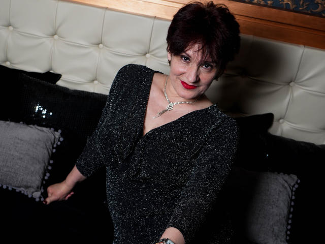 AlbertaAllen with short dark red hair, red lipstick, wearing a glittery black dress, sitting on a black couch with sequined and gray pillows, smiling at the camera.