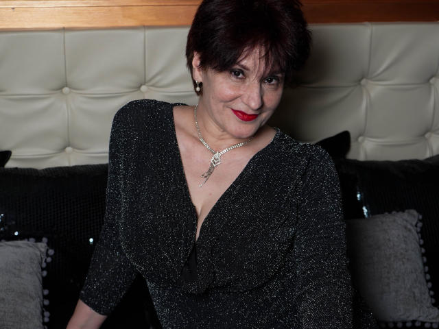 Alberta Allen, com cabelo curto e escuro, batom vermelho e um vestido preto brilhante, usando um colar, sentada em um sofá preto com almofadas, sorrindo para a câmera. Seus seios estão visíveis..