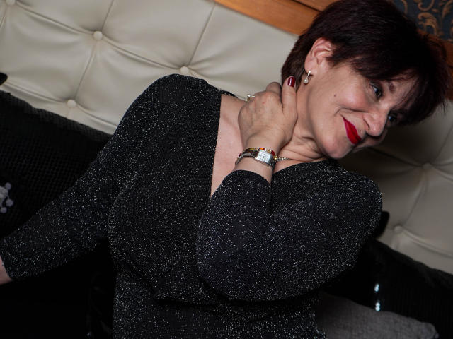 AlbertaAllen with short dark hair, red lipstick, black sparkly top, holding her neck, smiling, wearing a silver watch, earrings, and red nail polish, sitting on a black couch.