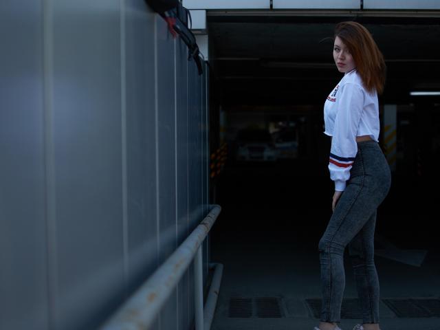 CuttieEyesX heeft rood haar, draagt een witte crop top en strakke grijze broek, en staat in een parkeergarage met een metalen muur aan de linkerkant en een donkere achtergrond..