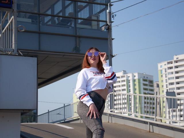 CuttieEyesX, com cabelo ruivo, vestindo uma blusa cropped branca com listras vermelhas e azuis, e calças cinzas justas, segurando óculos de sol, em frente a um prédio de vidro..