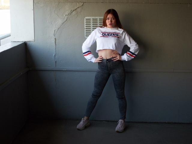 CuttieEyesX está de pie con las manos en las caderas, vistiendo una camiseta corta blanca de Queens que deja ver su abdomen, y unos vaqueros oscuros ajustados, con zapatillas grises, en una habitación con paredes grises..