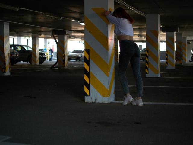 CuttieEyesX, vêtue d'un débardeur blanc et d'un pantalon moulant gris, appuyée contre un pilier rayé de jaune dans un parking sombre, porte des talons hauts blancs..