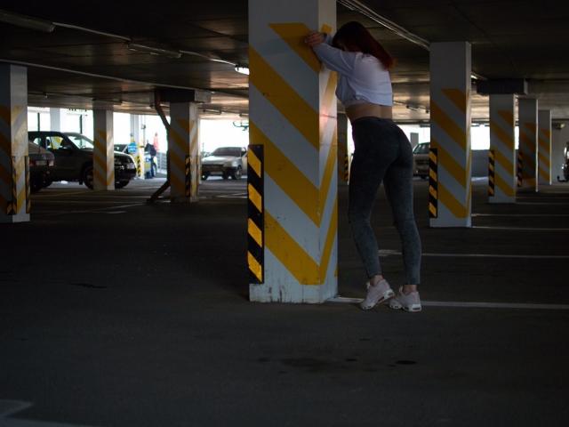 CuttieEyesX lehnt sich an eine gelb gestreifte Säule in einer dunklen Parkgarage, trägt ein weißes Crop-Top, graue Leggings und weiße High Heels und zeigt dabei ihren Hintern..