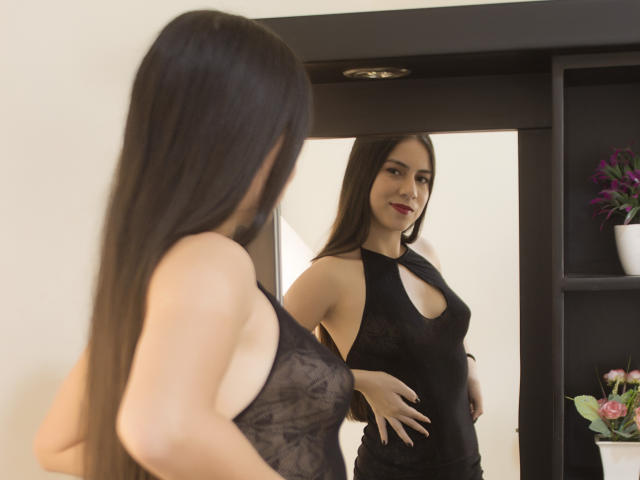 JuliaGarciax with long black hair, wearing a sheer black halter dress, standing in front of a mirror, smiling, showing her tits and small ass, reflecting in the mirror.