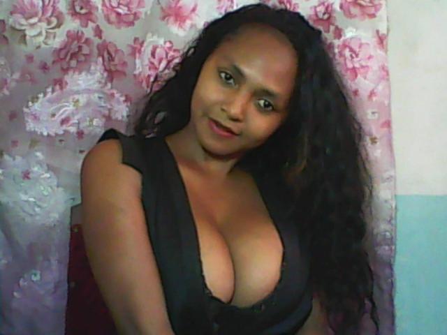 LeCorp with dark skin, curly black hair, wearing a black dress with deep cleavage, showing large boobs, smiling, standing against a floral-patterned wall.