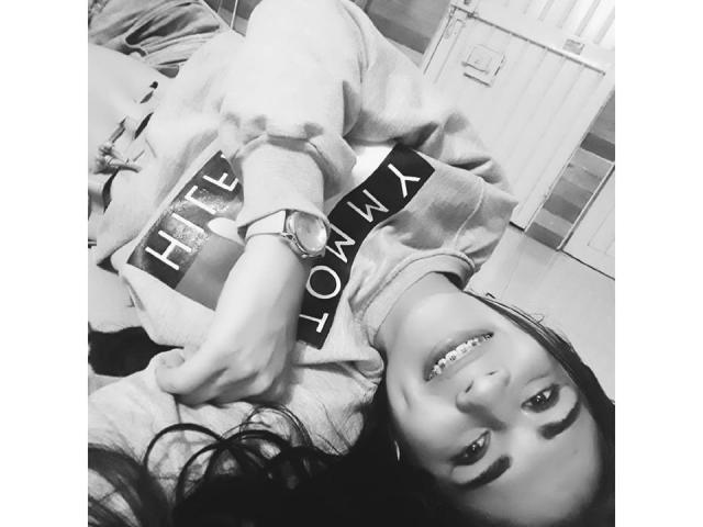 CristalHugeX acostada boca arriba, sonriendo a la cámara, con un reloj puesto, sosteniendo un libro en la mano, en una foto en blanco y negro, mostrando sus pechos..