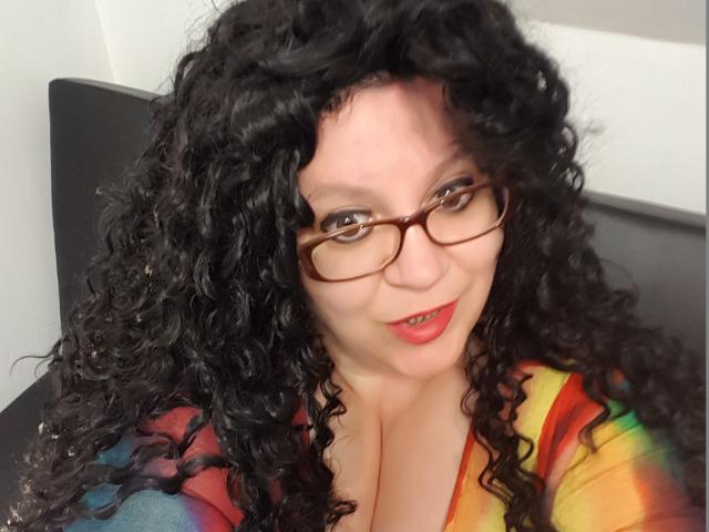 FoxDollBBW with curly black hair, glasses, and red lipstick, wearing a rainbow shirt, showing large boobs, smiling slightly, seated on a black chair.
