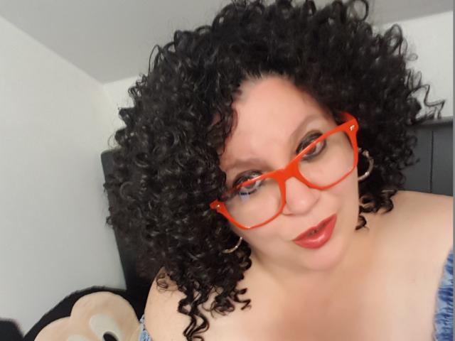 FoxDollBBW con capelli neri ricci, occhiali rossi e rossetto rosso, che indossa una maglietta blu a spalle scoperte e sorride alla fotocamera. Seno visibile, di medie dimensioni, pelle morbida..