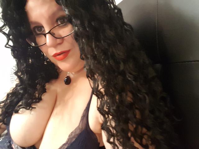 FoxDollBBW with curly black hair, glasses, red lipstick, and deep cleavage in a lace purple bra, wearing a black choker with a purple gem. Boobs.
