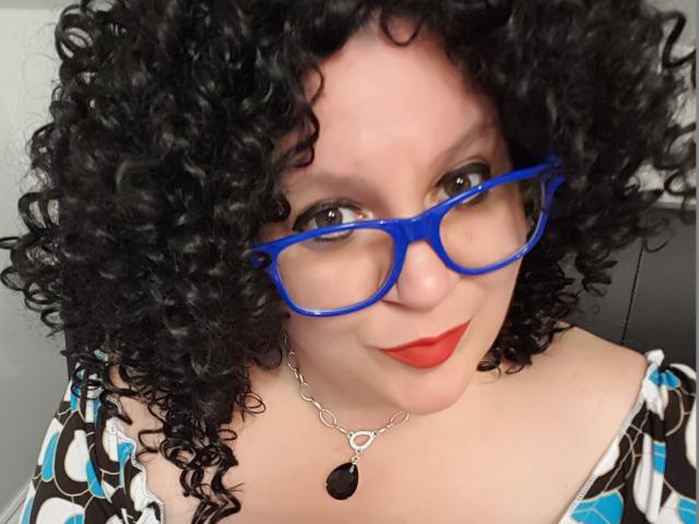 FoxDollBBW con capelli neri ricci, occhiali blu, rossetto rosso e una maglietta a fantasia, indossa una collana d'argento con una pietra nera, e guarda verso la fotocamera..
