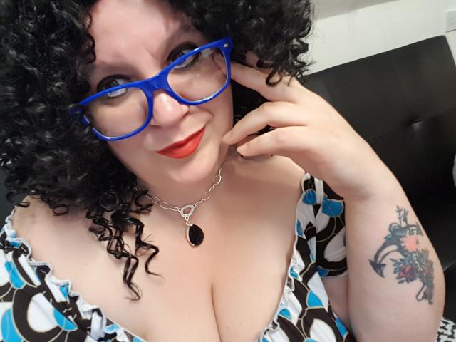 FoxDollBBW con capelli ricci, occhiali blu, rossetto rosso, scollatura, vestito bianco e blu a motivi, collana d'argento, tatuaggio sul braccio destro, che tocca il viso, sorridente, pelle pallida, seno di media grandezza..