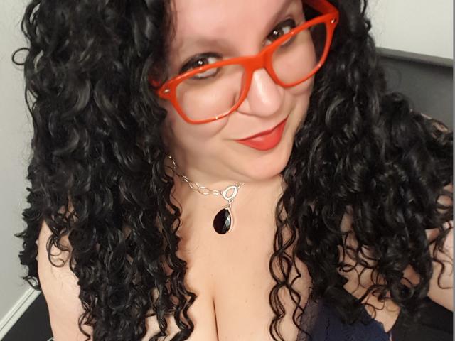 FoxDollBBW with curly black hair, red glasses, red lipstick, and a black necklace with a large black gem, showing her big boobs. She's smiling at the camera.