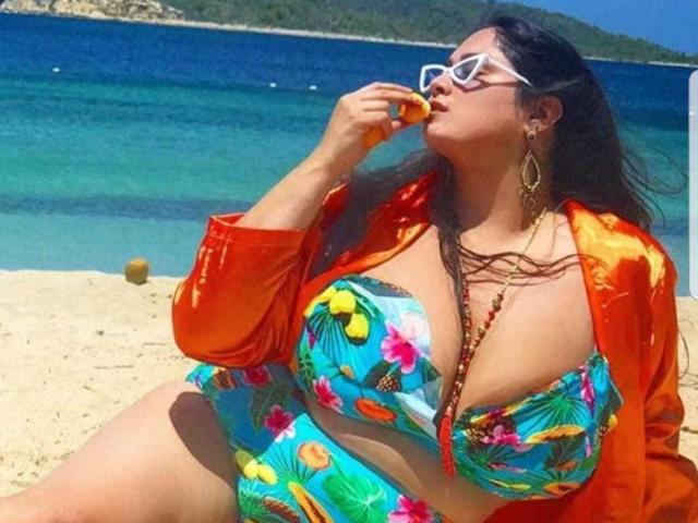 FoxDollBBW con grandi seni, in un bikini tropicale colorato, una maglietta arancione, occhiali da sole bianchi e orecchini lunghi, che mangia della frutta su una spiaggia assolata..