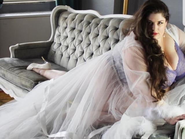 FoxDollBBW with long brown hair, purple lace bra, and white sheer dress, lying on a grey tufted sofa, looking seductively at the camera, showing her tits.