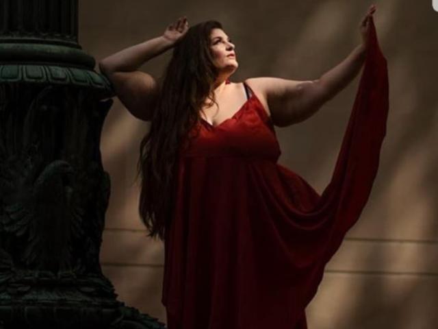 FoxDollBBW with long brown hair, wearing a red dress, holds it up, arms raised, standing beside a dark, ornate pillar, looking off to the side, in soft light.