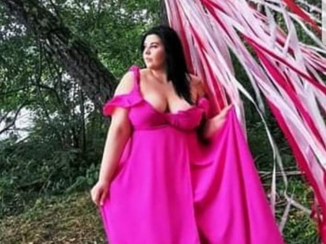 Una FoxDollBBW di taglia grande, con grandi seni, è in piedi in una foresta, indossa un abito rosa acceso, tiene in mano il tessuto, con nastri rossi e bianchi sullo sfondo..
