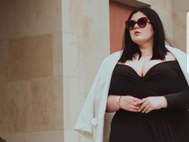 FoxDollBBW con capelli neri, occhiali da sole e rossetto rosso, indossa un abito nero scollato, una giacca bianca e un braccialetto, ed è in piedi contro un muro di pietra beige. Il suo seno è molto evidente..