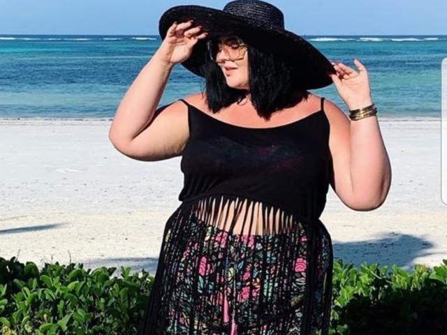 FoxDollBBW con caschetto nero, che indossa un grande cappello nero, occhiali, una maglia nera con frange, una gonna floreale e un braccialetto dorato, in piedi su una spiaggia, con il cappello in mano..