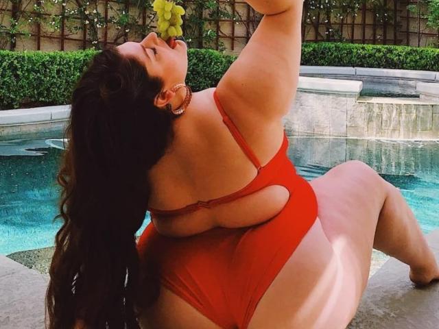 FoxDollBBW with large ass and big tits, sitting by the pool, wearing a red one-piece, holding a grape to her mouth, long dark hair, pool water in background.