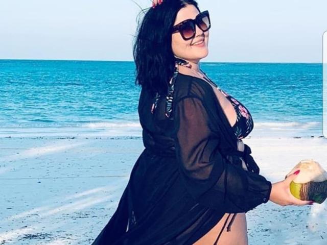 FoxDollBBW con capelli neri, occhiali da sole e un abito trasparente nero, che tiene un cocco, indossa un bikini floreale, mostrando seni grandi, in piedi su una spiaggia con oceano blu..