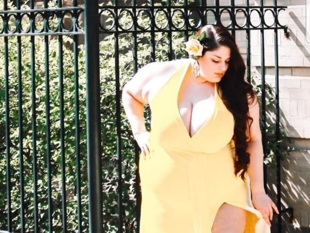FoxDollBBW with big tits, huge yellow dress, long black hair, yellow flower, standing by black iron gate, sunny, looking down, hand on thigh, green foliage in background.