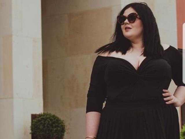 FoxDollBBW con i capelli neri, che indossa occhiali da sole, un abito nero con una spalla scoperta e mostra il suo seno prosperoso, è in piedi con una mano sul fianco, guardando in alto. Ha una figura formosa e una carnagione chiara..