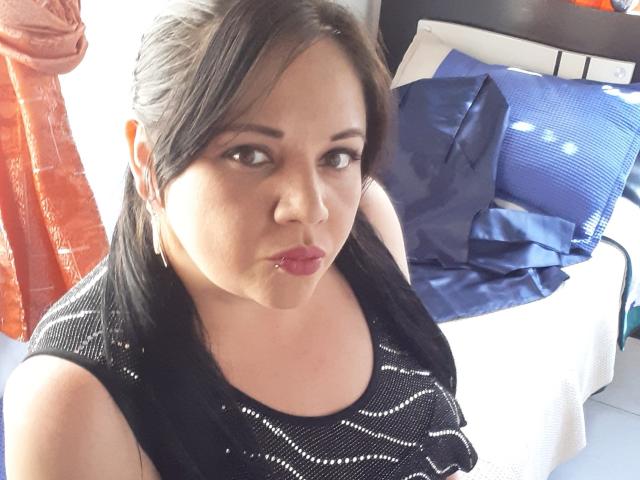 Alis69, com cabelo preto, vestindo uma blusa preta com estampas, lábios vermelhos e finos, e brincos, sentada em frente a uma cama com travesseiros azuis e laranjas..