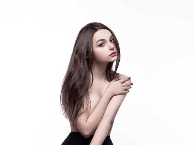 AnirieRozental with long brown hair, topless, covering small tits with one hand, black skirt, pale skin, red nail polish, white background, looking slightly to the right.