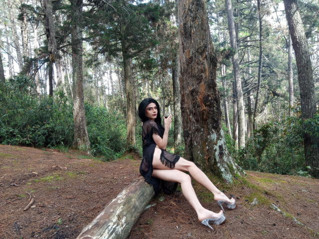 Rosariio with pale skin, black sheer dress, and clear heels, sitting against a tree in a forest, legs crossed, touching her chin, looking at the camera, showing her ass.