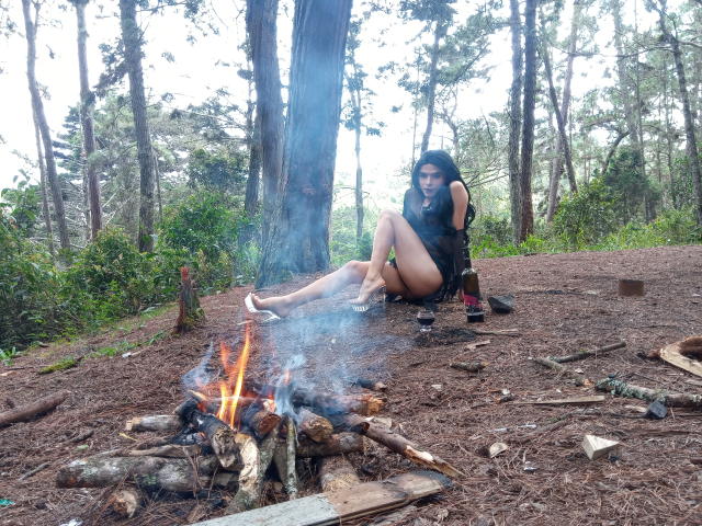 Rosariio with long black hair, sitting by a small fire in a forest, wearing a black top and no pants, legs up, surrounded by bottles and logs.