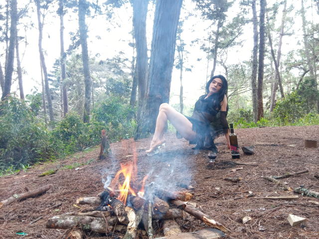 Rosariio mit langen schwarzen Haaren, einem grünen Oberteil und Shorts, sitzt am Feuer, die Beine angezogen, eine Flasche und ein Glas liegen auf dem Boden, im Hintergrund ein Wald, sie raucht, trägt Stiefel, das Gesäß ist sichtbar..