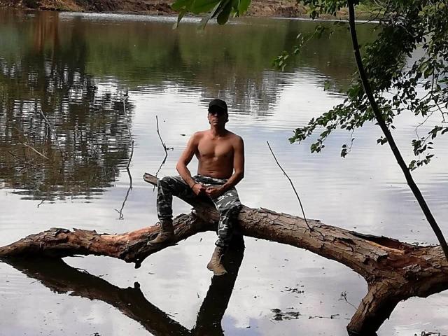 Un couple sauvage pratiquant le fétichisme des corps nus et musclés est assis sur un arbre tombé, portant un pantalon de camouflage et des bottes, au bord d'une rivière calme et paisible, avec des feuilles vertes au premier plan..