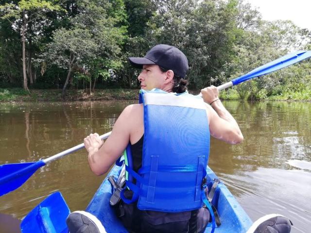 Fétiche de couple sauvage avec un gilet de sauvetage bleu, une casquette noire et une tresse, pagayant un kayak bleu sur une rivière boueuse, tenant une pagaie bleue, dos à la caméra, bras musclés..