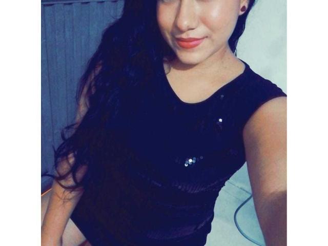 JadenaZir with long black hair, wearing a black sequin top, sitting, showing her face with orange lipstick, and slightly smiling. Her tits are visible through the top.
