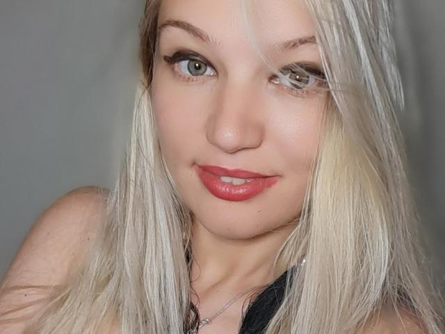 AlmaDeAngel69 has blonde hair, green eyes, and red lipstick. She's smiling with slightly parted lips, showing a hint of her teeth. Her makeup includes black eyeliner.