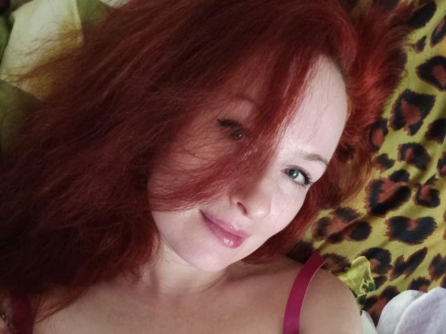 Marlene Davies, met rood haar, roze lippen en groene ogen, liggend op een kussen met luipaardprint, met een roze bandje om, lachend naar de camera..