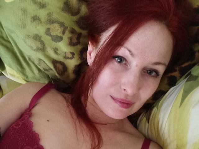 MarleneDavies has red hair, blue eyes, and is wearing a pink lace bra, lying on a leopard print pillow with a green pillow behind her.