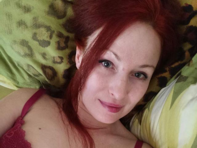 MarleneDavies with red hair, green eyes, and pink lipstick, lying on a leopard-print pillow, wearing a red lace bra, smiling softly at the camera.